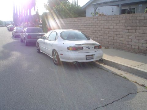 1993 Ford Taurus - A white sedan, likely a 1996 Ford Taurus, is parked on the street. Other vehicles are visible in the background.