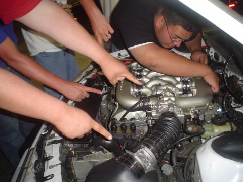 1993 Ford Taurus - Several people examine the engine bay, with fingers pointing towards the intake manifold and other engine components.