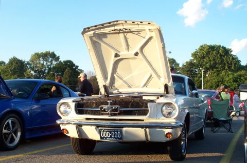 1998 Ford Crown Victoria - A white 1965 Ford Mustang is parked with its hood open at a car show, next to a blue modern car.
