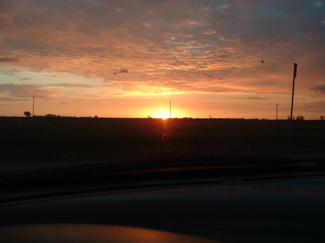 Sunrise over flat fields seen through a windshield with the dashboard in the foreground