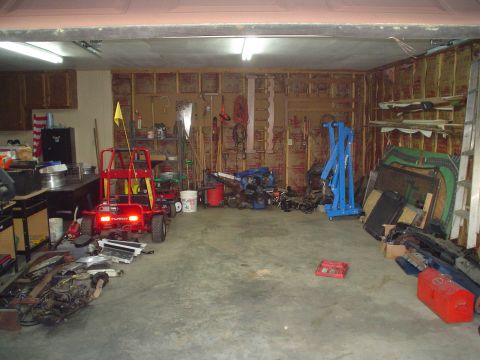 1981 Ford Bronco - A cluttered garage scene contains various tools, engine components, and a red go-kart, with unfinished walls and shelving.