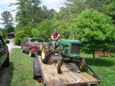 1981 Ford Bronco - A man drives a tractor loaded onto a trailer, which is being pulled by a pickup truck.