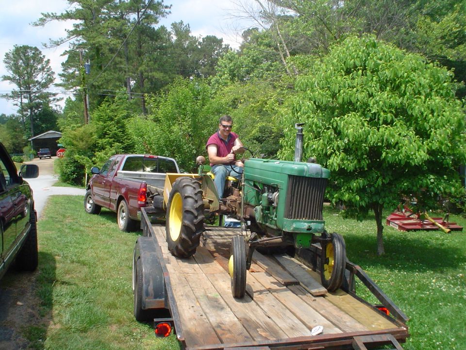 Green tractor on a wooden trailer being towed by a red pickup truck