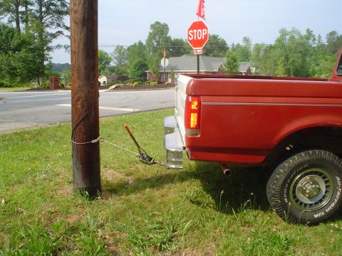 1981 Ford Bronco - Rear view of a red pickup truck with a tow strap connected to a utility pole, possibly preparing to move it.