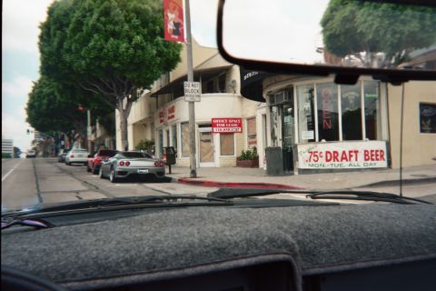 1990 Ford Bronco - Through a windshield, a city street corner shows a coffee shop and a hand-painted 75-cent draft beer sign with parked cars.
