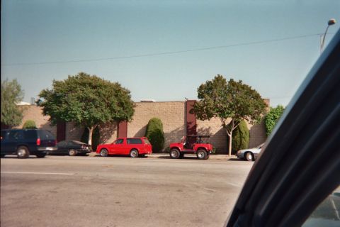1990 Ford Bronco - red SUV and a small red 4x4 buggy are parked along a street in front of a cinder-block wall and trees, shot from a car.