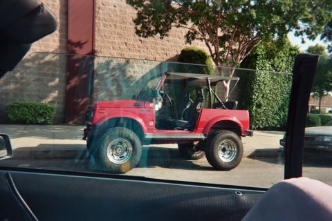 1990 Ford Bronco - small red lifted soft-top 4x4 SUV sits in a parking lot outside a brick building, reflected in the photographer's car window.