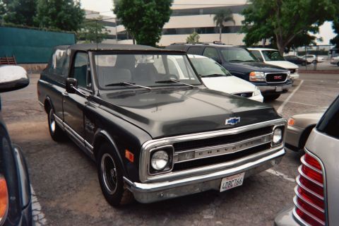 1990 Ford Bronco - dark green classic Chevrolet C10 pickup with a black top and bowtie grille badge is parked in a lot among modern cars.