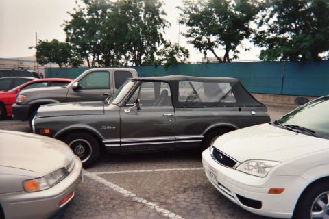 1990 Ford Bronco - gray vintage Chevrolet K5 Blazer with its soft top folded down is parked between a white car and a sedan in a paved lot.