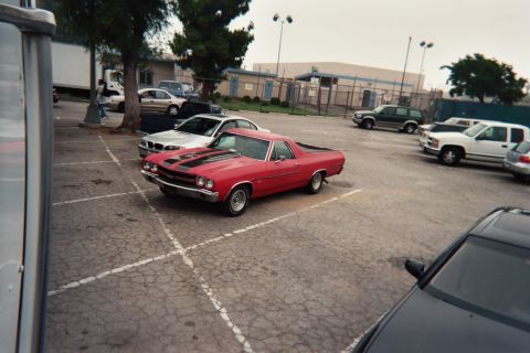 1990 Ford Bronco - red Chevrolet El Camino with black hood stripes is parked at an angle in a cracked parking lot near an industrial building.