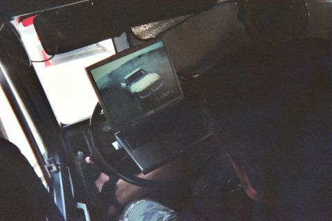 1990 Ford Bronco - laptop is mounted over a steering wheel inside a dim vehicle cabin, its screen showing a photo of a light-colored vehicle.