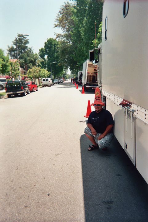 1990 Ford Bronco - man wearing a foam hat crouches beside a large white enclosed trailer on a sunny tree-lined street with traffic cones.
