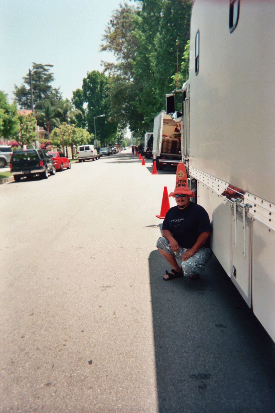 Man in a foam hat crouching beside a large white enclosed trailer on a tree-lined street