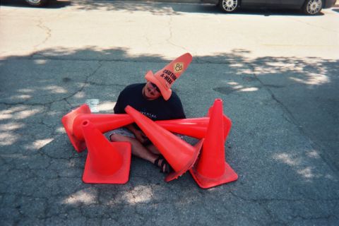 1990 Ford Bronco - grinning man sits cross-legged on pavement surrounded by orange traffic cones, balancing a cone on his head.