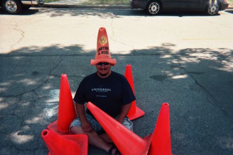1990 Ford Bronco - man wearing a 'Caution Ahead' traffic-cone novelty hat sits surrounded by orange traffic cones on cracked asphalt.
