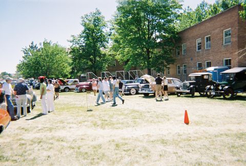 1972 Ford Ranchero - People gather around vintage cars displayed on a grassy field outside a brick building.