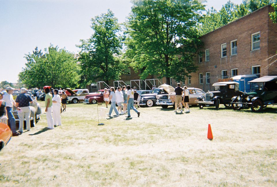 Classic car show on grass lawn beside brick building with spectators and a black Model T