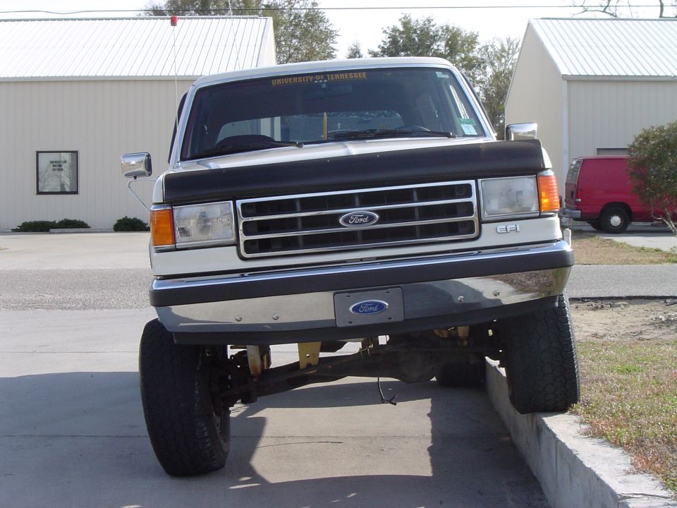 Front view of a lifted white Ford F-Series truck with a black hood protector. University of Tennessee sticker on windshield. Red van in background