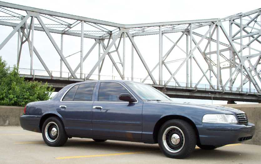 Dark blue Ford Crown Victoria sedan parked in front of a steel truss bridge