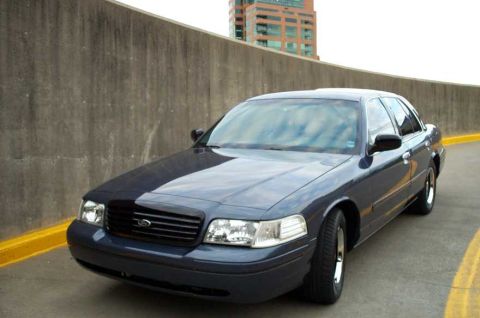 1998 Ford Crown Victoria - A dark blue Ford Crown Victoria is parked near a concrete barrier with a yellow line on the road.