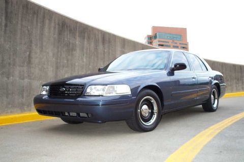1998 Ford Crown Victoria - A dark blue sedan is parked on a road with a concrete wall and a building in the background.