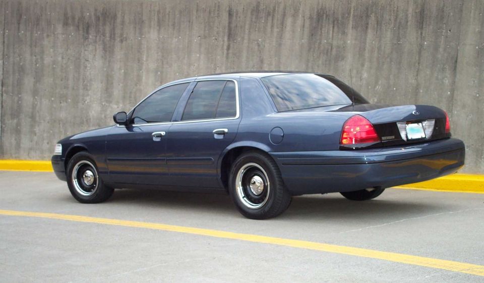 Rear three-quarter view of dark blue Crown Victoria on steel wheels beside concrete wall