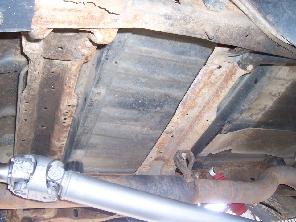 Underside view of a truck's frame, showing the driveshaft and exhaust system. Rust is visible on the frame components