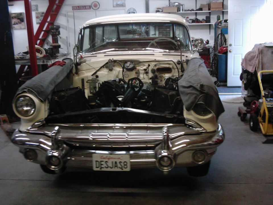 Front view of a classic car with its hood open, showing the engine bay. The car is in a garage with tools and shelves in the background. License plate reads 'DESJAS8'