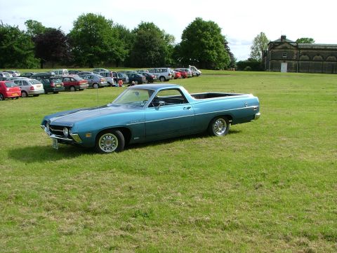 1970 Ford Ranchero - A blue 1970 Ford Ranchero is parked on a grassy field, with a building and a parking lot full of cars in the background.