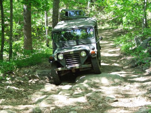 1996 Ford Bronco - An olive drab jeep navigates a challenging rocky trail in a wooded area, with a second vehicle following closely behind.