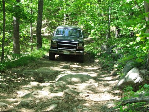 1996 Ford Bronco - A dark green SUV drives through a challenging, rocky path surrounded by lush green trees.