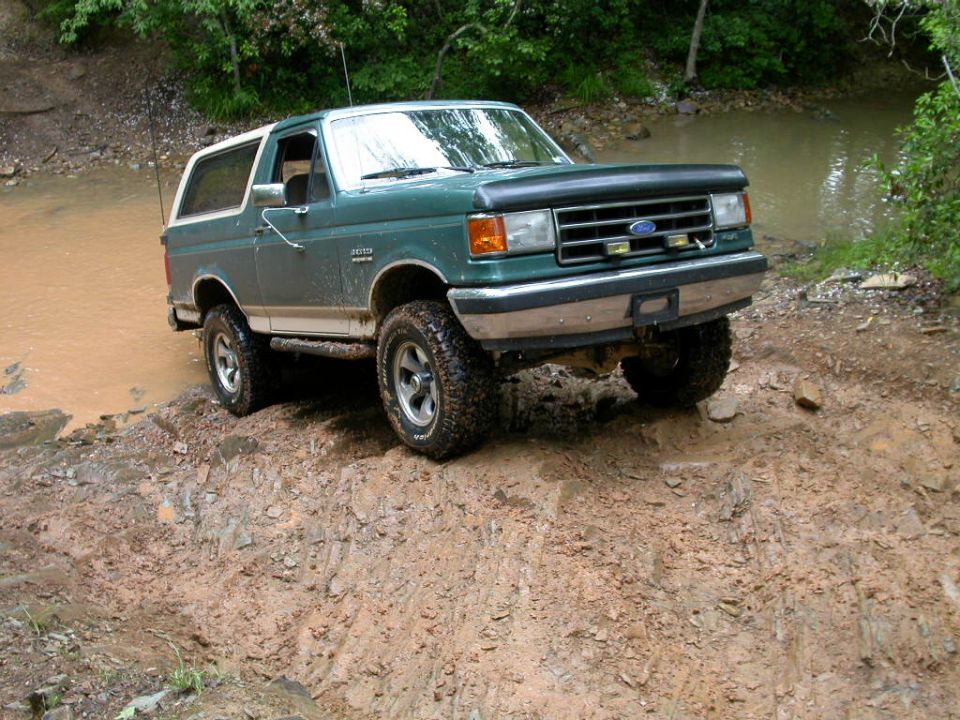 Green and tan Ford Bronco in muddy water crossing on a rocky creek bank