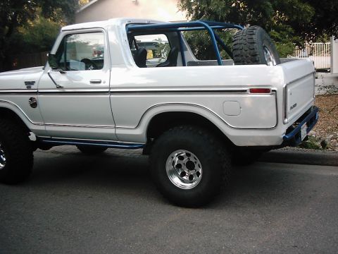 1978 Ford Bronco - A white 1978 Ford Bronco with the top removed, shown from the driver's side rear 3/4 angle, featuring a blue roll bar, rear-mounted spare t…
