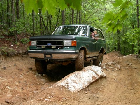 1990 Ford Bronco - A green Ford Bronco tackles a challenging off-road trail in a wooded area.