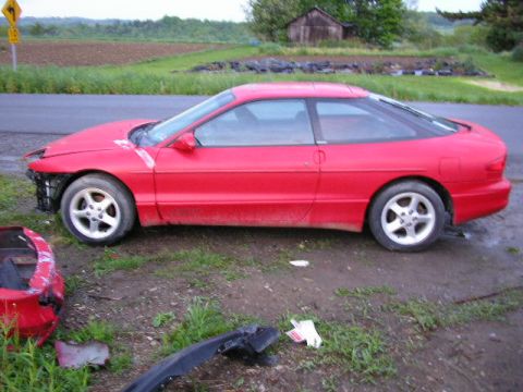 1994 Ford Probe - A damaged red coupe sits on the side of the road with debris scattered around it.
