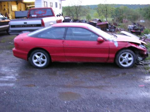 1994 Ford Probe - A red sports coupe sits damaged in a yard next to a pickup truck and other debris.