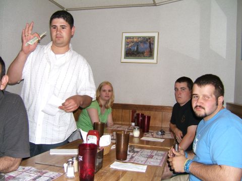 1998 Ford Taurus - A group of people are seated at tables in a diner, with one man standing and holding papers.
