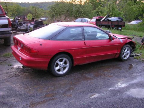 1994 Ford Probe - A red coupe sits damaged in a junkyard with other vehicles and debris in the background.