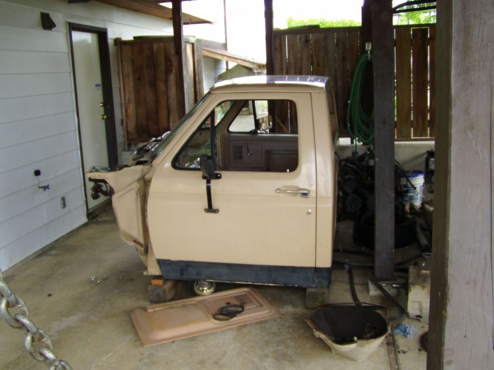 Tan Ford truck cab off the frame on blocks in a carport, door panel removed on ground
