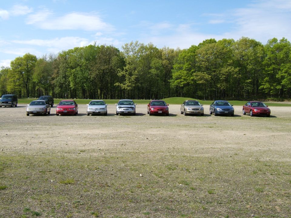 several sedans parked in a gravel lot with trees in the background