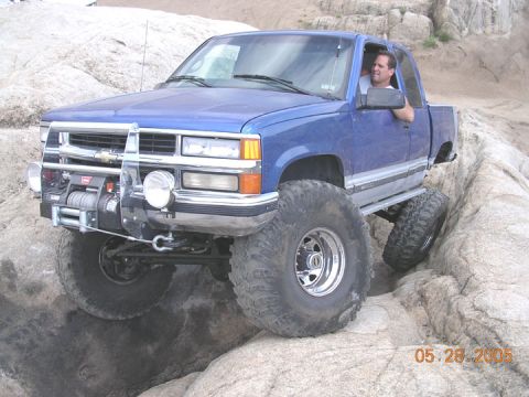 1994 Ford Bronco - A blue pickup truck is shown off-roading on a rocky surface, with one wheel lifted.