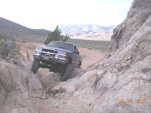 1994 Ford Bronco - A blue pickup truck is shown off-roading through a rocky landscape, with mountains in the background.