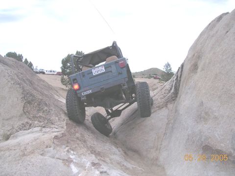 1994 Ford Bronco - A blue Jeep Wrangler navigates a rocky off-road trail, showing its rear and lifted suspension.