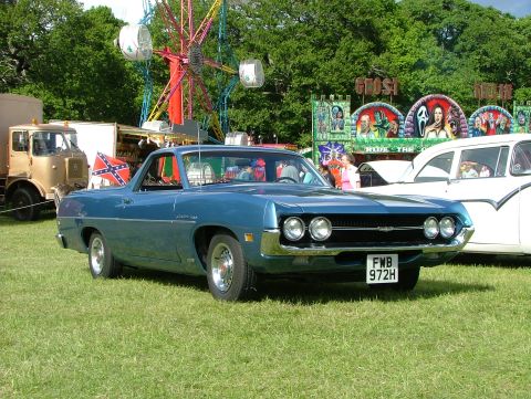 1970 Ford Ranchero - A blue 1970 Ford Ranchero is seen from the front, parked on a grassy field with carnival attractions behind it.