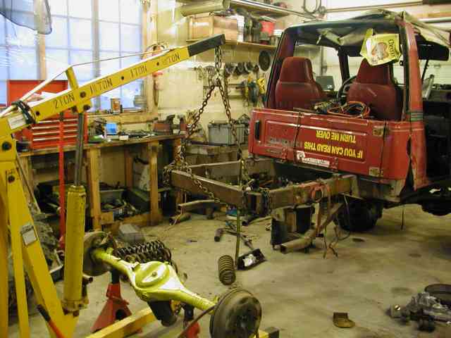 Red compact 4x4 body lifted off its frame by a yellow engine hoist in a home shop