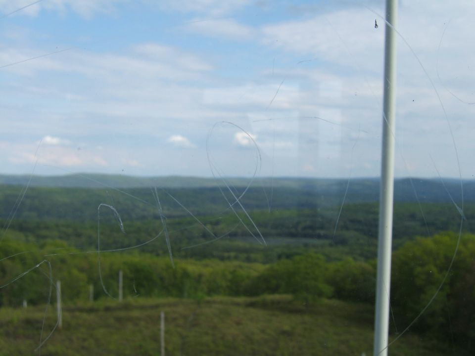View of green hills and blue sky with clouds through a window with many scratches. A white pole is visible on the right