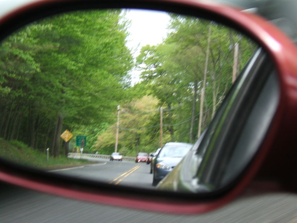 Side view mirror reflection shows a road with trees and multiple cars driving
