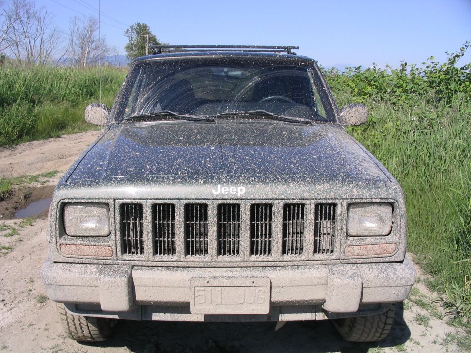 Front view of a muddy 2000 Jeep Cherokee parked on a dirt road with tall grass on either side
