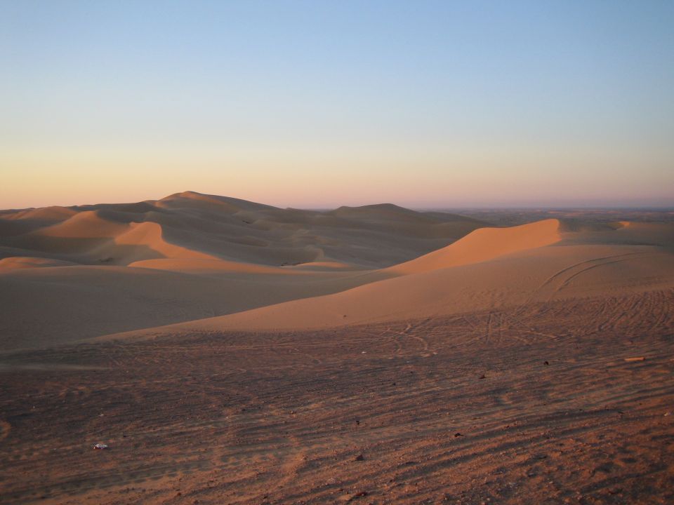 Desert sand dunes with tire tracks under a gradient sky at sunset