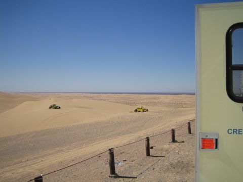 1968 Ford Mustang - Two dune buggies traverse sandy terrain under a vast blue sky, with a portion of a light-colored structure visible on the right.
